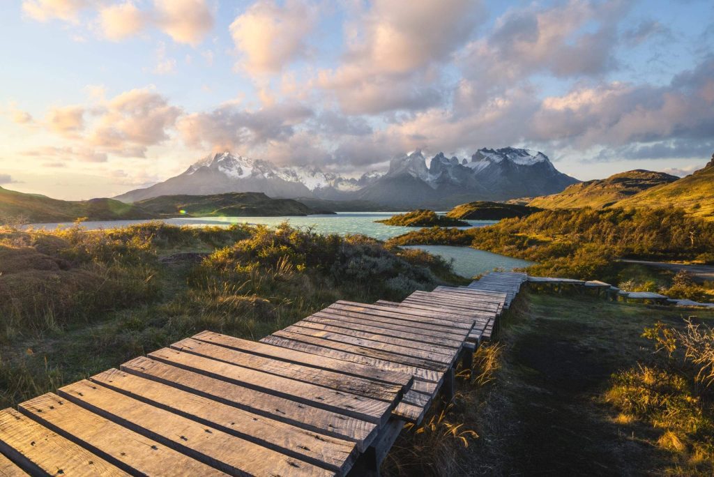 Wooden pathway leading to a serene lake with mountains in the background under a vibrant sunset sky with fluffy clouds.