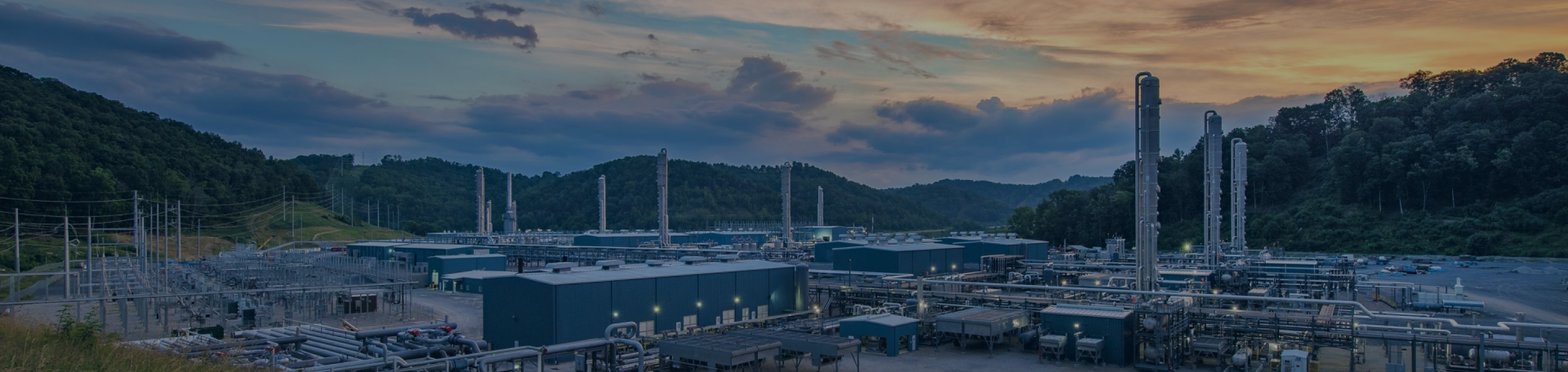 An industrial facility nestled in a hilly landscape at sunset. The facility consists of large structures and equipment spread out over a wide area, with tall, cylindrical towers rising into the sky.