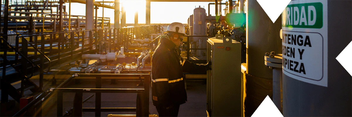 Worker operating machinery in an industrial plant at sunset, ensuring safety compliance with displayed signs in Spanish.